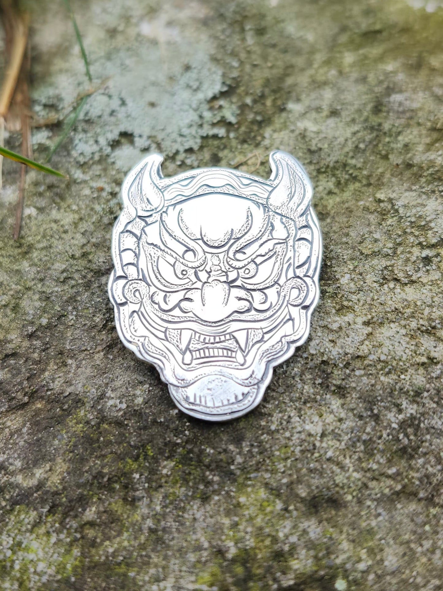 Hand-engraved sterling silver Oni mask pendant photographed on a textured moss-covered stone background.