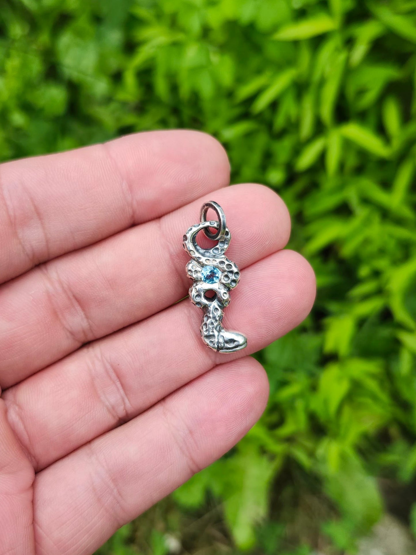 Hand holding the silver Serpentine Alchemy pendant with a vivid blue topaz gemstone, set against a background of greenery