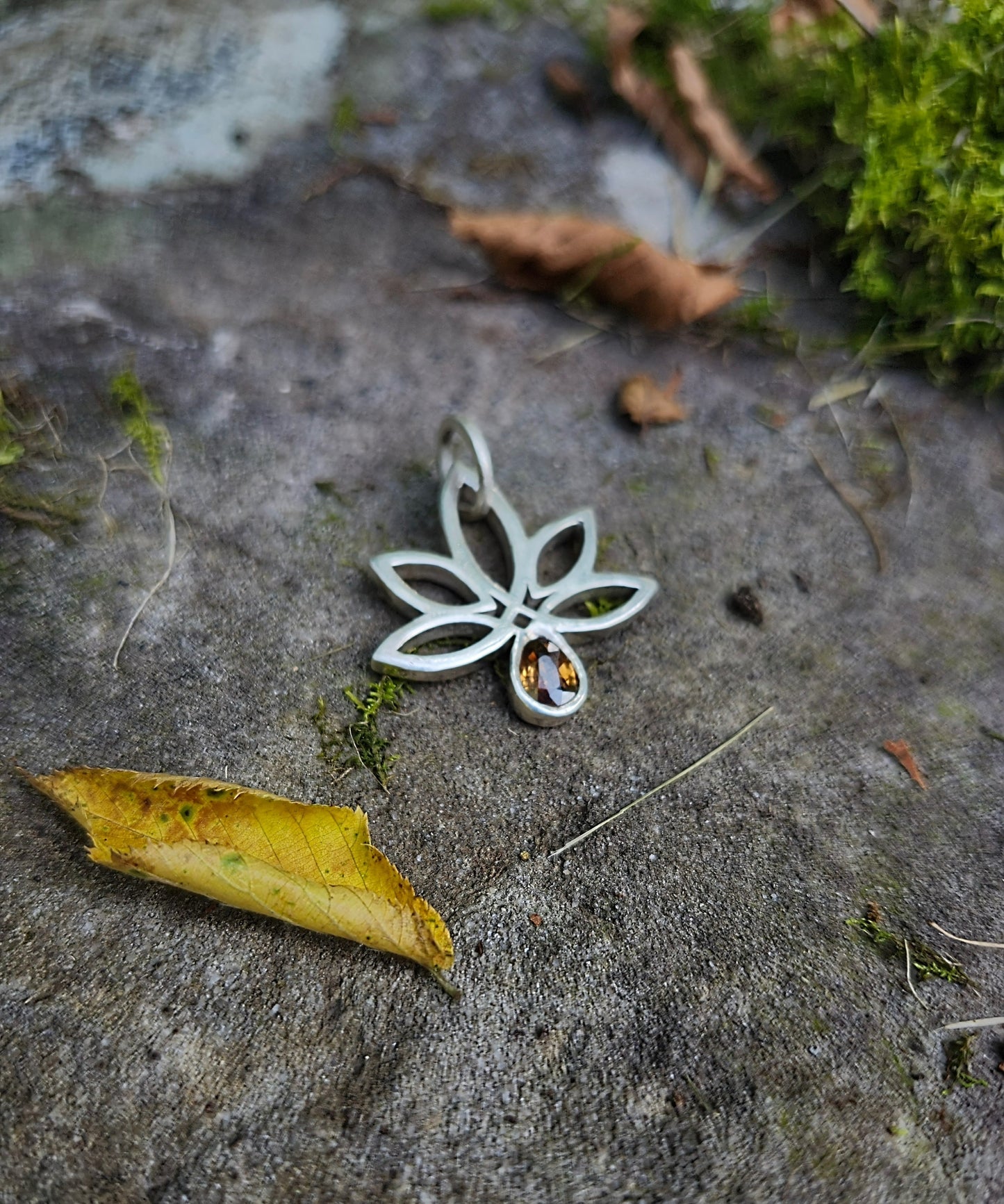 Silver flower-shaped pendant with a gemstone on a stone surface with moss and leaves.