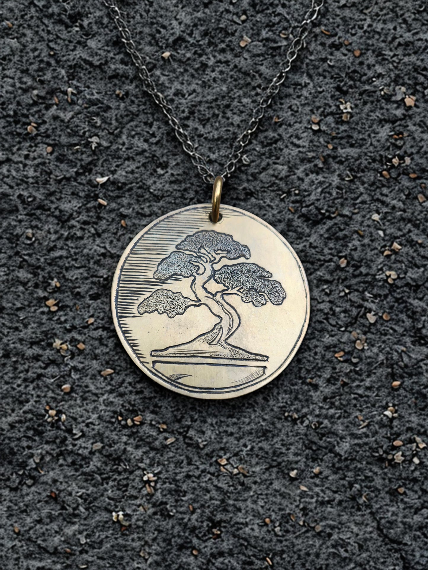Front of hand-engraved bonsai medallion pendant displayed on textured black stone background.