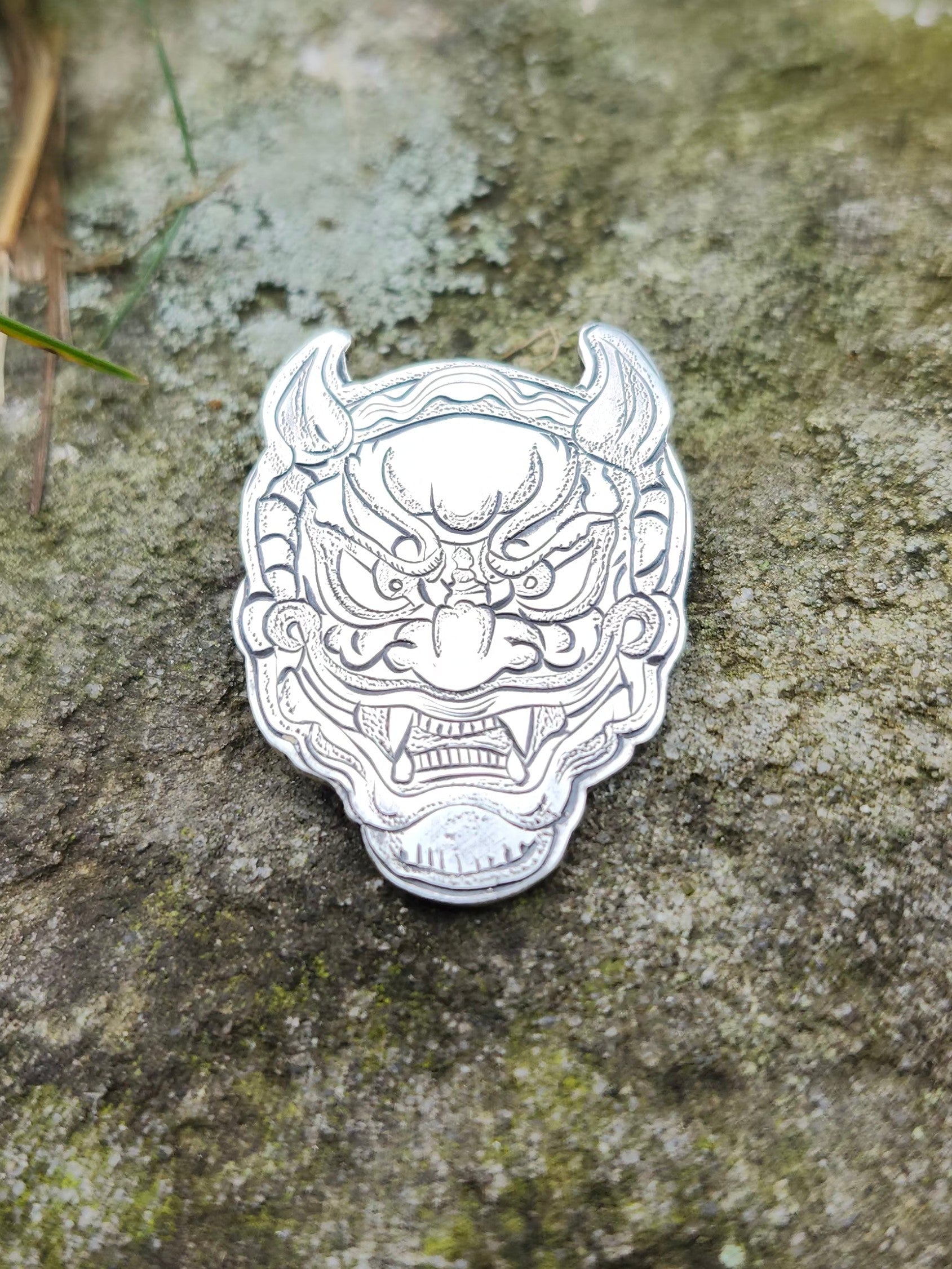 Hand-engraved sterling silver Oni mask pendant photographed on a textured moss-covered stone background.