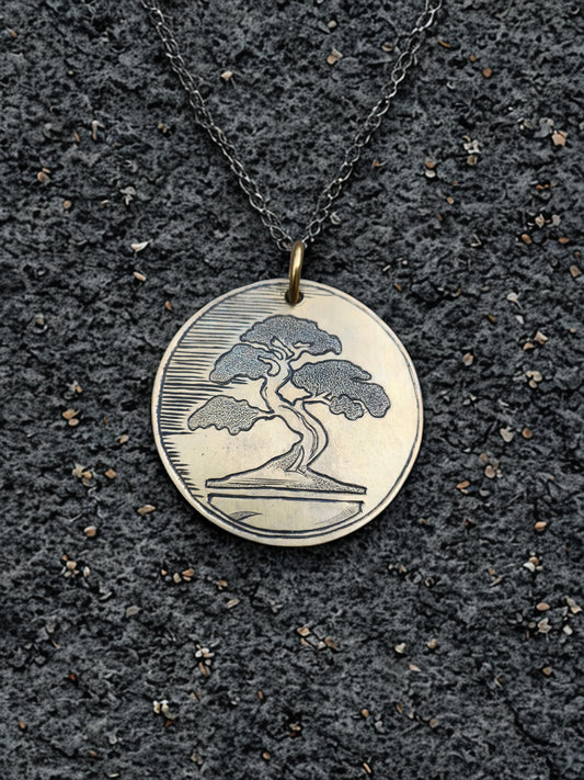 Front of hand-engraved bonsai medallion pendant displayed on textured black stone background.